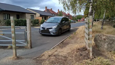 A grey car is pictured parked on a pavement next to wide gap framed by a metal gate and wooden fence on a residential street. There are brick houses on one side and grass and trees on the other.