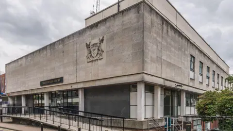 The outside of St Albans Magistrates' Court. It is a grey concrete building, in a box shape, with a coat of arms on the side and black metal ramps leading up to the building. There are windows and an entrance, and there is a tree on the right. 