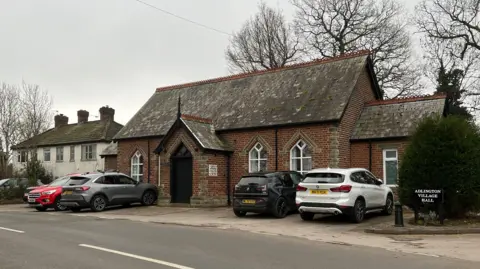 A view of Adlington VIllage Hall. It is an old redbrick building and has five cars parked outside.