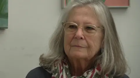 Susanna Rook wears glasses, a dark coloured top and a patterned scarf around her neck. She is seated and there is some artwork behind her.