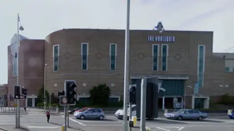 Google Two-storey brick building which curves round to the right in line with the road. It has tall narrow windows on the first floor and "THE HARLEQUIN" in 20s-style lettering towards the top above two of the windows. There are cars on the road in front of the building and a set of traffic light.