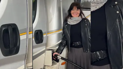 A woman with long dark hair, wearing a white scarf and a leather jacket fills up the fuel of the camper van.