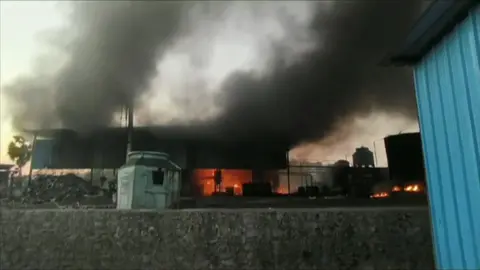 A still from footage of the aftermath of an explosion at a pyrolysis plant in India, showing a copper-blur structure in the foreground resembling a storage tank and a low-rise shed behind it which is ablaze, with thick black smoke rising into the sky. Under part of the shed, which is exposed at the sides, is a large, black pile of used tyres.