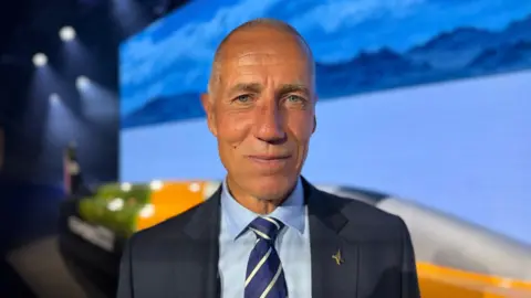 An man stands in front of a yellow car and a background.
