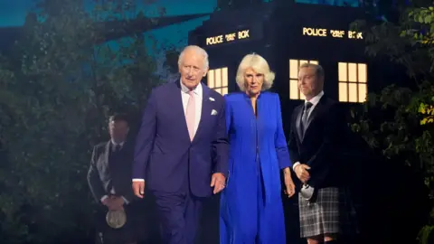 The King and Queen are walking in front of a Tardis. Sir Chris Hoy is standing behind them. 