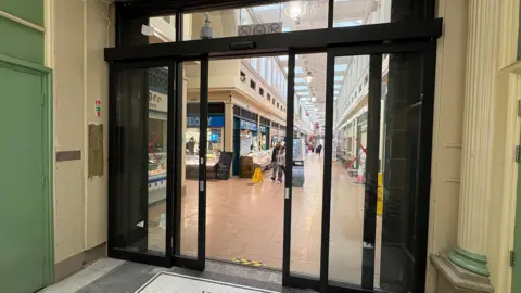 Newcastle City Council View of a sliding glass door, partially opened, and leading onto the arcade of an indoor market stretching on into the distance, with a few people strolling past the stalls.