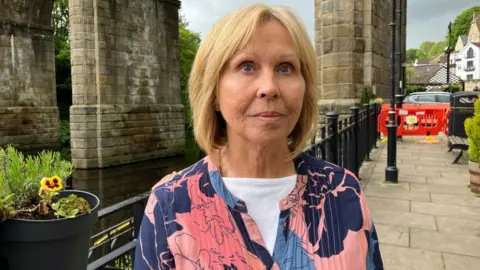 BBC/Seb Cheer A woman wearing a pink, purple and blue top looks into the camera lens with a neutral expression on her face.