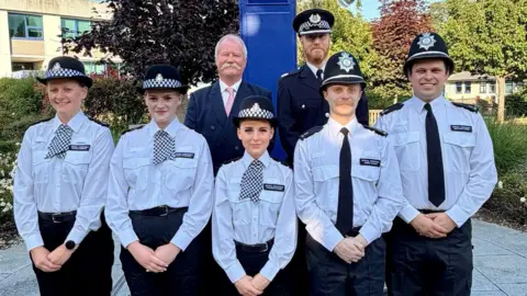 Five police officers stand in a row with their hands clasped before them. They are all wearing white shirts, black police hats and black trousers. Two men stand behind them, one in a suit with a pink tie and the other in formal police uniform. 
They are standing outside with greenery behind them. 