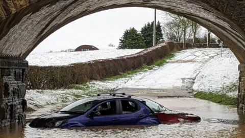 Two cars - one blue and one red - stuck in flooding under a bridge. Snowy fields and a road can be seen in the background.