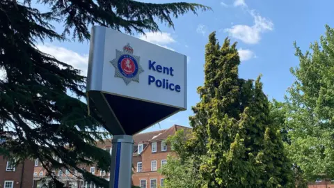 A sign which reads Kent Police. There is a large red building behind the sign and trees in front of the building.