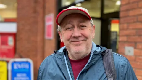 Andrew Twigg stands close to the doorway of the Post Office. He is wearing a turquoise waterproof jacket, white baseball cap with a red rim, and has a rucksack strap over his left shoulder