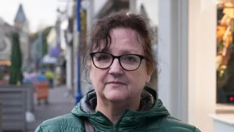 Lesley is pictured on the high street outside shops. She is wearing black glasses with her brown hair tied up. She has a green puffer jacket coat on.