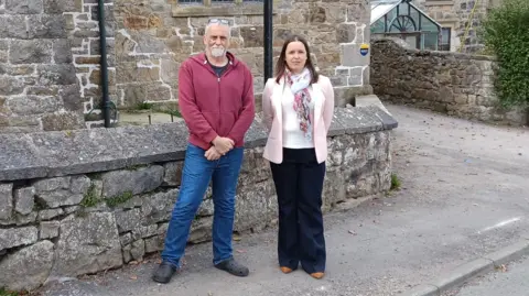 A man and a woman stood on the pavement outside a stone house.