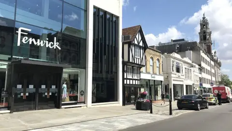 Martin Giles/BBC The exterior of Fenwick, as seen from Colchester High Street. The building has a mostly glass facade with the word Fenwick in front in white letters. There are cars parked in front of the building, including a police car.