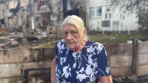 Hazel Tompkins is wearing a blue and white floral-patterned top stands in the foreground of a fire-damaged residential area. Behind the person are heavily charred homes, blackened walls and debris, with signs of extensive fire damage to the properties and surrounding gardens. The image is focused on the person, while the burnt buildings in the background are slightly blurred, highlighting the scale of destruction caused by the blaze.