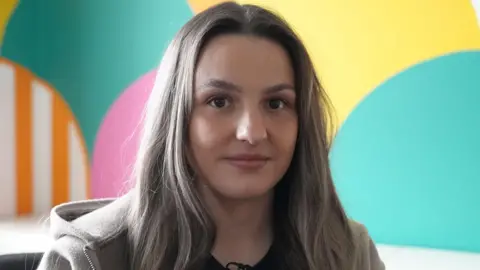 BBC Alaw looking at the camera smiling. She wears a grey jumper, black t-shirt and has long silver hair. 