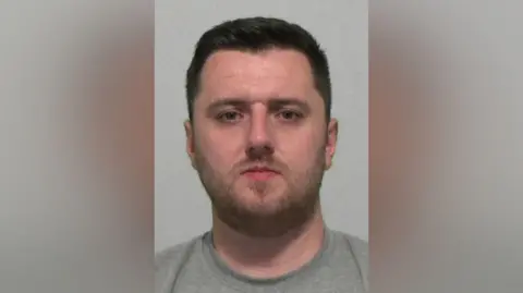 Mugshot of Callaghan. He has a large head and small mouth, with short dark hair and stubble. He is wearing a grey sweatshirt.
