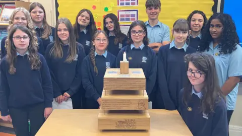 Thirteen students wearing blue high school jumpers stand around a table. They are all looking directly at the camera. On the table is a wooden structure made of three layers with a candle on top.