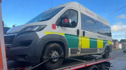 Merlin Brick A grey van that says "ambulance patient transport". It has green and yellow squared on it, and a sliding door. It is strapped to a recovery vehicle trailer