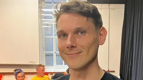 A man in a black T-shirt and with a gold necklace, stood in front of a creative arts class.