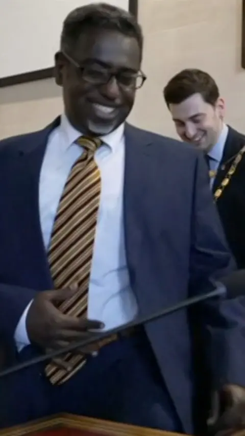 Yassin Mohamud is standing and smiling. He is wearing a blue suit, white shirt and a yellow and claret tie. He is inside a council chamber and the former Lord Mayor is in the background, he is also smiling.