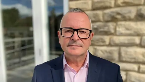 BBC/SAM READ Head and shoulder shot of Labor MP Lee Barron, wearing glasses, looking at the camera, standing in front of a stone wall and door