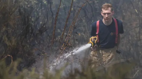 A male firefighter in a navy blue branded T-shirt, and tan brown trousers, points a hose with water coming out at dry grass.