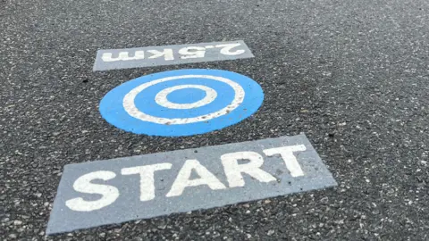 There are three parts to the marker which is on a tarmac path. The middle bit has five blue and white concentric circles, one block to the left of it reads: 2.5km and the other to the right reads: START.