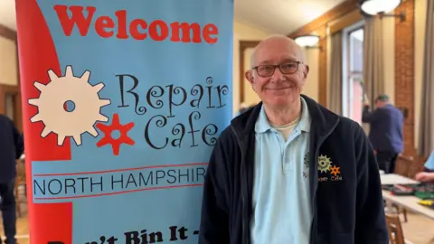 A man with white hair and glasses, wearing a navy fleece and blue polo shirt, both embroidered with a "repair cafe" logo. He is stood next to a blue and red banner which reads "welcome, repair cafe North Hampshire".