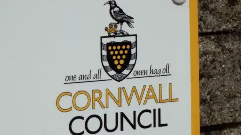 BBC White and yellow sign reads "One and all Cornwall Council". The sign is on a brick wall.