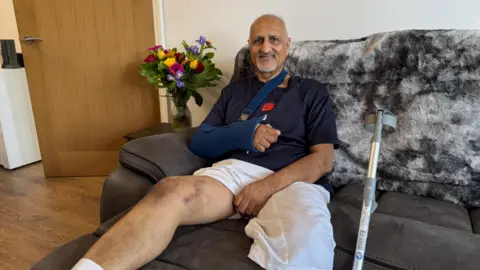 A man with short grey hair and a beard sitting on a dark grey sofa with a dark blue shirt and one bare leg up on a stool with a silver crutch beside him