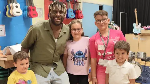 Contributed Smith and Matilda smile at the camera along with her three siblings. Smith is wearing a green shirt and jeans. They are standing in a room with a drum kit in the background. 