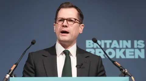Robert Jenrick speaks into a microphone from behind a podium, in front of a blue background with the slogan "Britain is broken".
