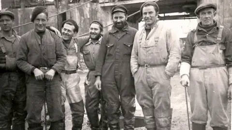 A group of seven whalers posing for the camera in a black and white picture. All are looking at the camera except the man in waders third from the left, who is staring at a man in overalls in a hat. The man on the near right is wearing waders and is leaning on a stick.