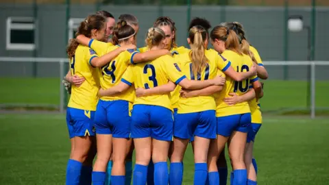 GT Photography A picture of a group of women footballers in blue and yellow kit huddled on a pitch.