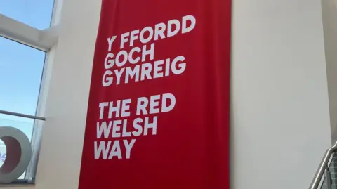 A red banner with "The Red Welsh Way" in English and Welsh at the front of Venue Cymru, with the blue skies over Llandudno beaming through a window to the left.