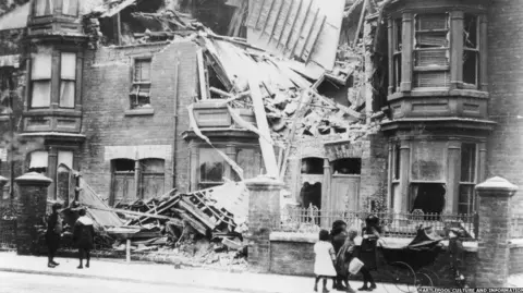 Hartlepool Museum and Gallery Service A black and white photograph of two groups of children looking at destroyed houses in a Hartlepool street. One of them is leaning on a pram.