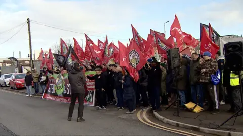 Unite the union members stand on the kerbside of a street in Birmingham. Flags are held aloft and a banner is placed in front of the picket line.