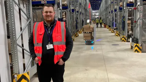 Sam Clarke has short dark hair and a ginger beard. He is wearing a hi vis jacket and is standing in a long corridor between enormous shelves.