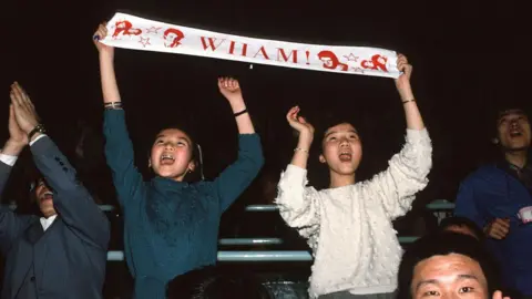 Getty Images Audience members cheer during a Wham! concert, with two fans holding a white scarf, bearing the band's name and images in red, above their heads. 