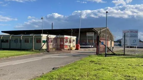 An exterior image of a second Atlas Leisure Homes site. The large warehouse stands behind a chain link fence and red coloured gates. The building is bathed in sunshine with blue skies and patchy cloud.