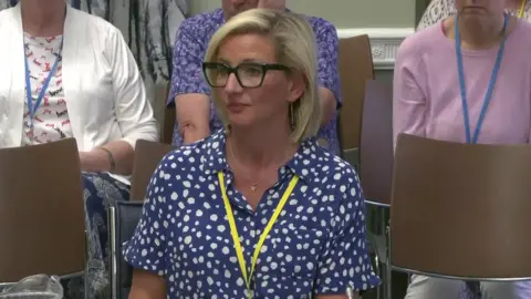 A screen grab of Roz McFeeters. She is sitting down and has short blonde hair and wearing black glasses. She has a blue top on that has white spots. She is wearing  bright yellow lanyard around her neck. 
