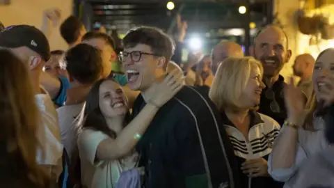A lively crowd gathers in a dimly lit pub, with people celebrating.