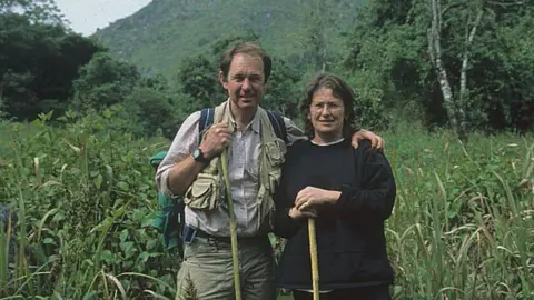 Bleddyn and Sue Wynn Jones in tall grass with trees and mountains in the background