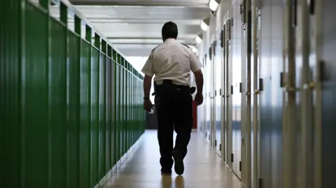 A stock photo of a male prison guard with his back to the camera, walking down a prison wing. The doors are on the right and green railings are to the left.