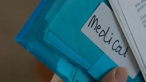 The image shows a close-up of a hand holding a bundle of papers and plastic document wallets. Several blue folders are visible, one with a handwritten label reading "Medical". On top of the folders is an out-of-focus printed letter, suggesting official correspondence.