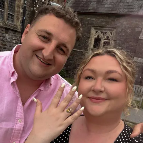 Kayley Stead Kayley showing off her engagement ring with hand up wearing a black dress with white polka dots and Richard with arm over her shoulder in pink shirt