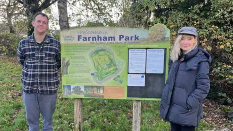A man stood to one side of an information sign in a park and a woman stood to the right. They are both looking into the camera. Behind them is grass and trees