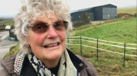 Rosemary Reakes wearing a brown coat and glasses. She has curly white hair and in the background is a farm.