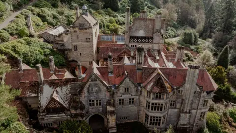 An aeriel view of the roof of the grand Cragside house. The mansion has multiple chimneys poking out of its red roof. The building is grey stone and surrounded by lush trees.